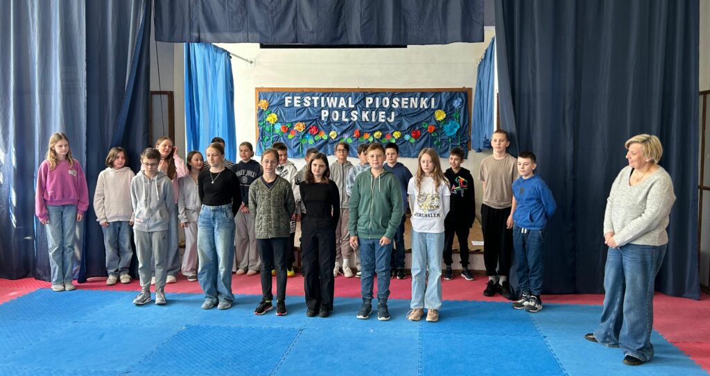 Uczniowie klasy 5b z nauczycielką współorganizującą śpiewają piosenkę. Jedni są uśmiechnięci, inni skupieni na aktywności. Stoją na scenie w dwóch rzędach, po obu stronach wiszą granatowe zasłony, w tle widnieje tablica z napisem Festiwal Piosenki Polskiej i kolorowymi kwiatami.