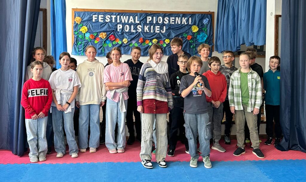 Uczniowie klasy 5c śpiewają piosenkę. Jedni są uśmiechnięci, inni skupieni na aktywności. Stoją na scenie w trzech rzędach: w pierwszym dziewczynka i chłopiec trzymający mikrofon, w drugim - osiem osób, w trzecim rzędzie- dziesięć osób. Po obu stronach wiszą granatowe zasłony, w tle widnieje tablica z napisem Festiwal Piosenki Polskiej i kolorowymi kwiatami.