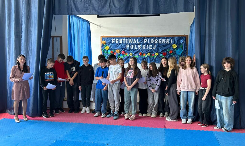 Uczniowie klasy 6b z wychowawczynią śpiewają piosenkę. Niektórzy trzymają w ręku kartkę z tekstem. Jedni są uśmiechnięci, inni skupieni na aktywności. Stoją na scenie w dwóch rzędach, po obu stronach wiszą granatowe zasłony, w tle widnieje tablica z napisem Festiwal Piosenki Polskiej i kolorowymi kwiatami.