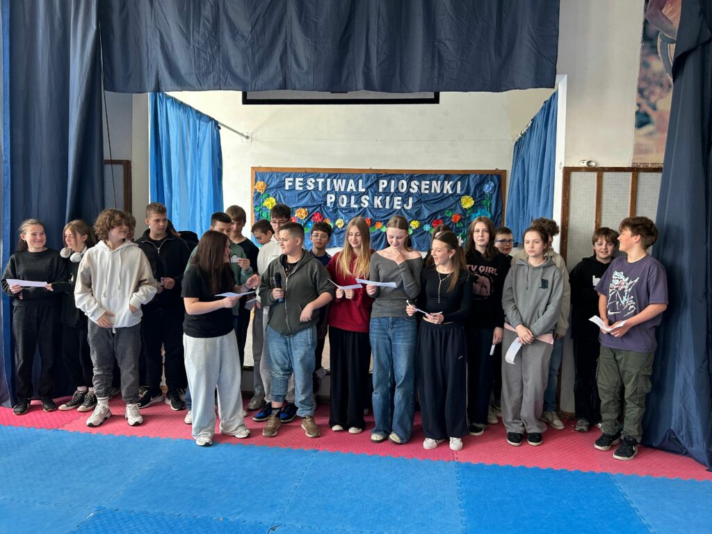 Uczniowie klasy 7b śpiewają piosenkę. Niektórzy trzymają w ręku kartkę z tekstem. Jedni są uśmiechnięci, inni skupieni na aktywności. Stoją na scenie, po obu stronach wiszą granatowe zasłony, w tle widnieje tablica z napisem Festiwal Piosenki Polskiej i kolorowymi kwiatami.