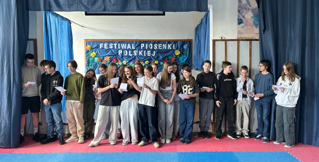 Uczniowie klasy 7c śpiewają piosenkę. Niektórzy trzymają w ręku kartkę z tekstem. Jedni są uśmiechnięci, inni skupieni na aktywności. Stoją na scenie, po obu stronach wiszą granatowe zasłony, w tle widnieje tablica z napisem Festiwal Piosenki Polskiej i kolorowymi kwiatami.