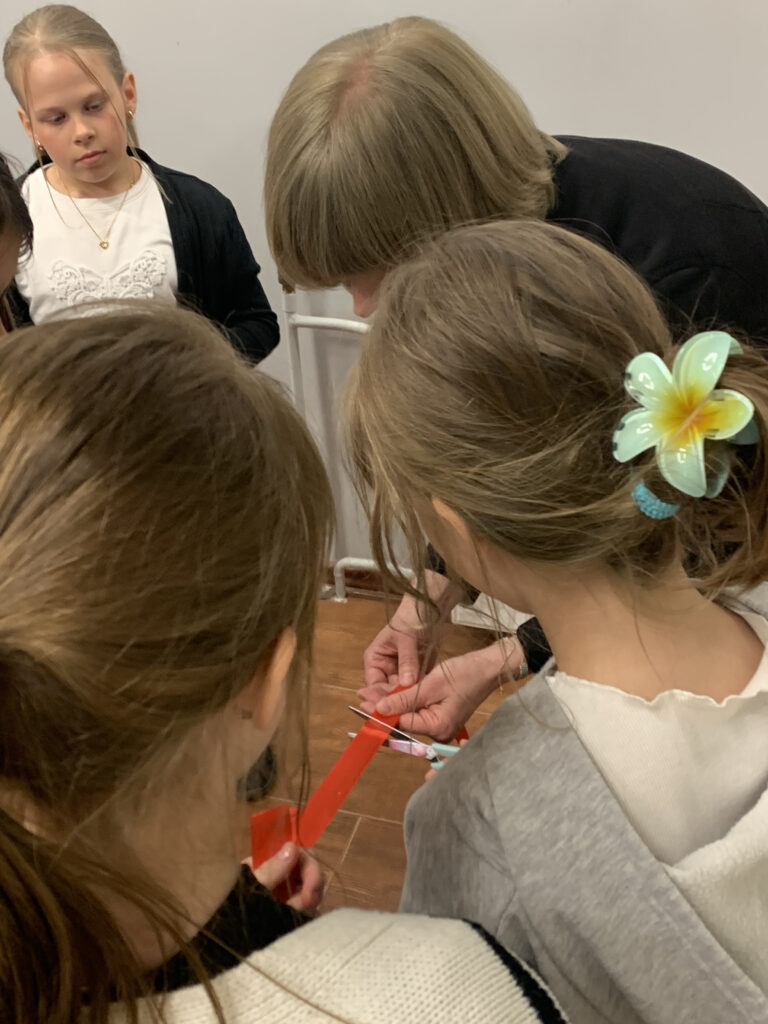 głowy trzech osób pochylone. W środku widoczna jest wstążka trzymana przez nie i ręce z nożyczkami przecinające wstążkę