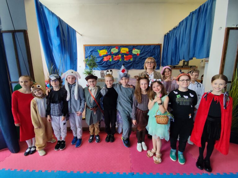 Zdjęcie przedstawia grupę uczniów klasy 2c wraz z wychowawczynią po zakończonym przedstawieniu „Zdrowy Czerwony Kapturek”. Dzieci stoją w dwóch rzędach, uśmiechnięte i dumne ze swojego występu. Ubrane są w kolorowe kostiumy przedstawiające postacie z bajki oraz różne zwierzęta i owady, takie jak wilk, myśliwy, babcia, Czerwony Kapturek, zając, sarenka, wróżka i inne. W tle widoczna jest dekoracja sceniczna z hasłami projektu „Zdrowo jem, więcej wiem”, a całość otaczają niebieskie kurtyny.