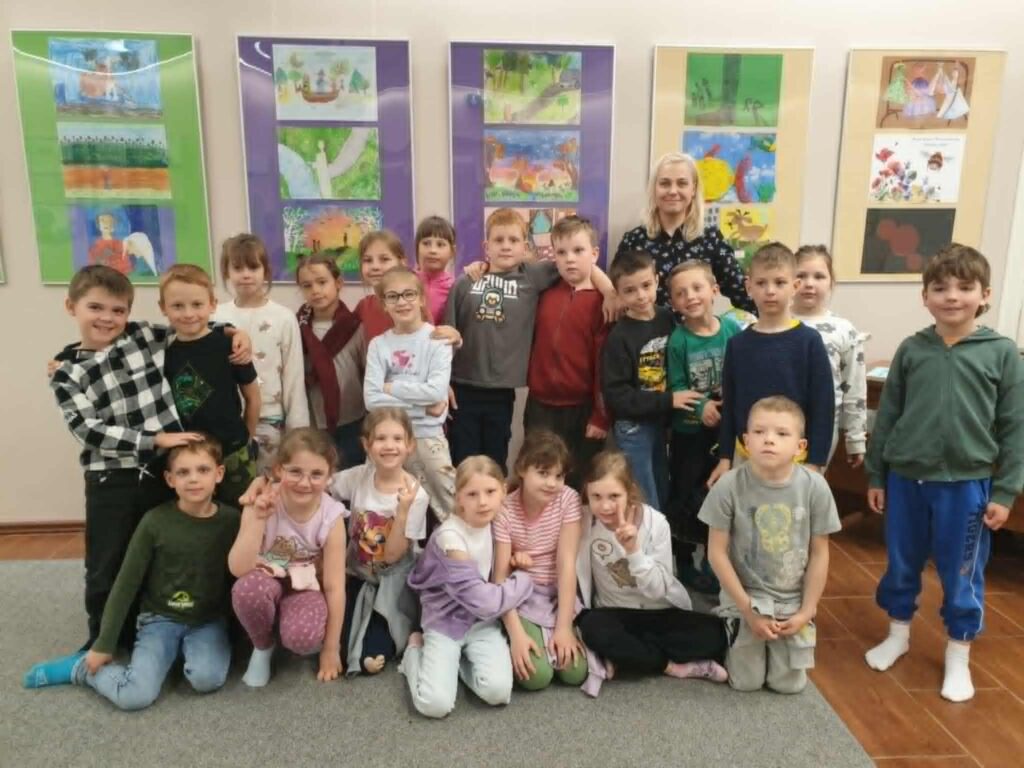 2. Na zdjęciu dzieci stoją w kole, trzymając się za ręce, a w tle na ścianie wyświetlony jest obraz Ziemi z zaznaczonym ruchem obrotowym. Nauczycielka po lewej stronie trzyma hula-hop, które najwyraźniej przekazywane jest po okręgu jako część zabawy edukacyjnej. W sali panuje półmrok, co podkreśla efekt projekcji, a ściany zdobią kolorowe prace plastyczne.