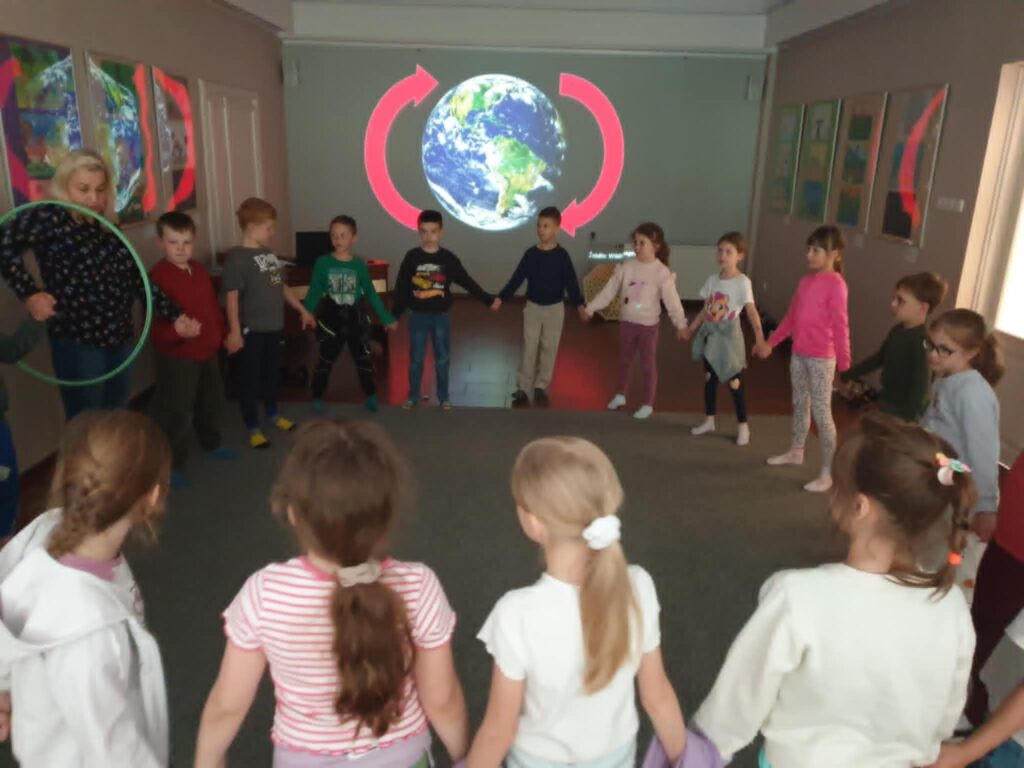 3. Na zdjęciu widać grupę dzieci siedzących w kręgu na wykładzinie w pomieszczeniu w bibliotece. W centrum uwagi znajduje się chłopiec trzymający skrzyneczkę, obok którego siedzi nauczycielka i kilku innych uczniów. Dzieci wyglądają na zaangażowane i zaciekawione tym, co pokazuje chłopiec. Na ścianie za nimi widoczne są kolorowe prace plastyczne, wykonane przez dzieci. Atmosfera na zdjęciu wydaje się być ciepła i edukacyjna.