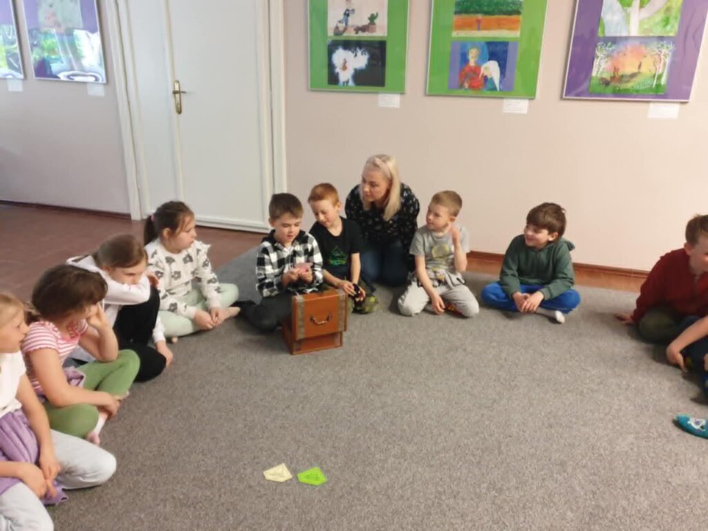 3. Na zdjęciu widać grupę dzieci siedzących w kręgu na wykładzinie w pomieszczeniu w bibliotece. W centrum uwagi znajduje się chłopiec trzymający skrzyneczkę, obok którego siedzi nauczycielka i kilku innych uczniów. Dzieci wyglądają na zaangażowane i zaciekawione tym, co pokazuje chłopiec. Na ścianie za nimi widoczne są kolorowe prace plastyczne, wykonane przez dzieci. Atmosfera na zdjęciu wydaje się być ciepła i edukacyjna.