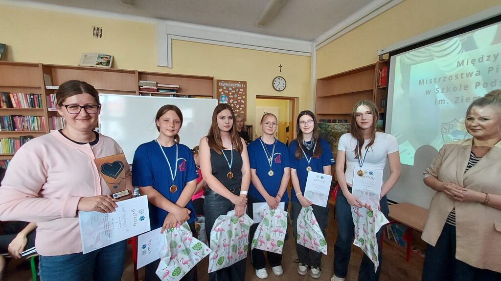 Zdjęcie przedstawia grupę dziewcząt stojących na tle ściany książek tablicy kropka dziewczęta mają w dłoniach dyplomy reklamówki z nagrodami na szyjach wiszą medale w kolorze brązowym. Obok dziewcząt z jednej strony stoi kobieta trzymająca w ręku statuetkę i dyplom kobieta ma okulary. Z drugiej strony stoi kobieta z rękoma złożonymi pod piersiami. W tle widać plakat.