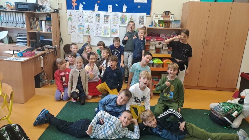 Zdjęcie przedstawia grupę chłopców i dziewcząt siedzących leżących bądź klęczących na podłodze część jest na zielonym dywanie część na żółtej podłodze bez dywanu. Stoją oni na tle rysunków przedstawiających Muminki. Wokół znajdują się szafy biurko półka z zabawkami.