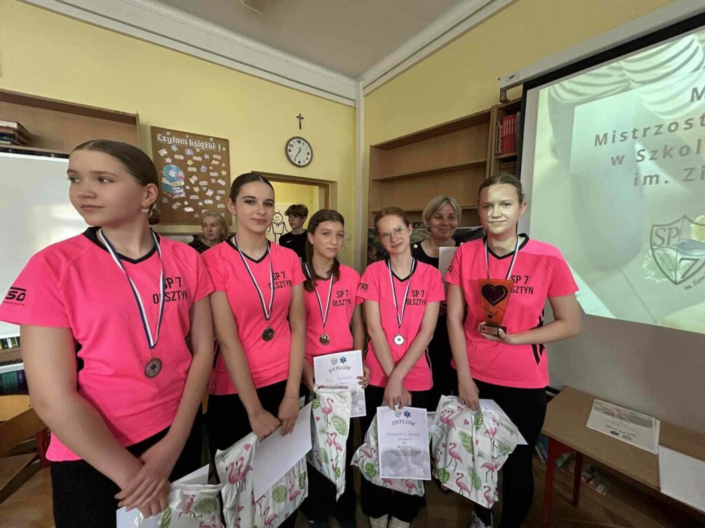 Zdjęcie przedstawia grupę pięciu dziewcząt trzymających w rękach dyplomy oraz reklamówki z nagrodami. Na szyjach dziewczęta mają medale w kolorze srebrnym. Ubrana są w różowe koszulki czarne spodenki. Jedna z dziewczyn trzyma w ręku statuetkę z czarnym serduszkiem. Dziewczyna bliżej fotografa jest odwrócona Patrz w głąb sali. Z tyłu za grupą dziewczyn stoi uśmiechnięta kobieta. W tle widać chłopca otwarte drzwi kobieta ściana z gazetką zegar.