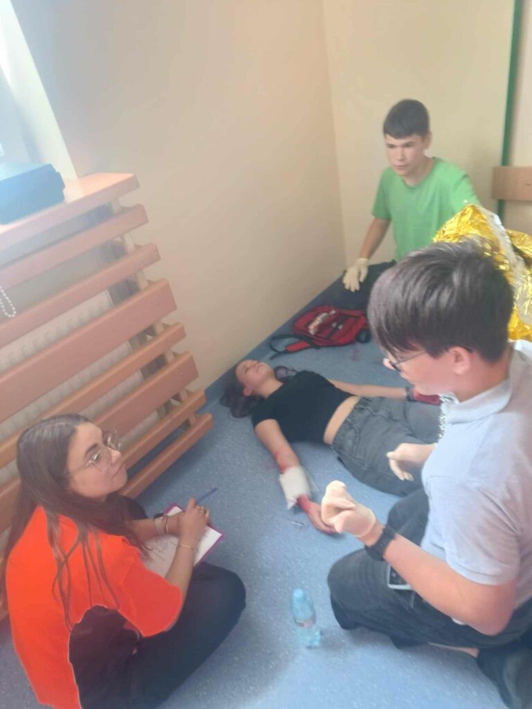 Zdjęcie przedstawia dziewczyny leżącą na podłodze z bandażem na rynku i zakrwawioną ręką. Obok dziewczyny znajduje się dwóch chłopców i jeden trzyma medyczny koc drugi mówi do kobiety która siedzi obok grupy. Kobieta ma pomarańczową koszulkę w dłoniach trzyma długopis i podkładkę siedzi po turecku. Chłopcy mają nałożone rękawiczki.