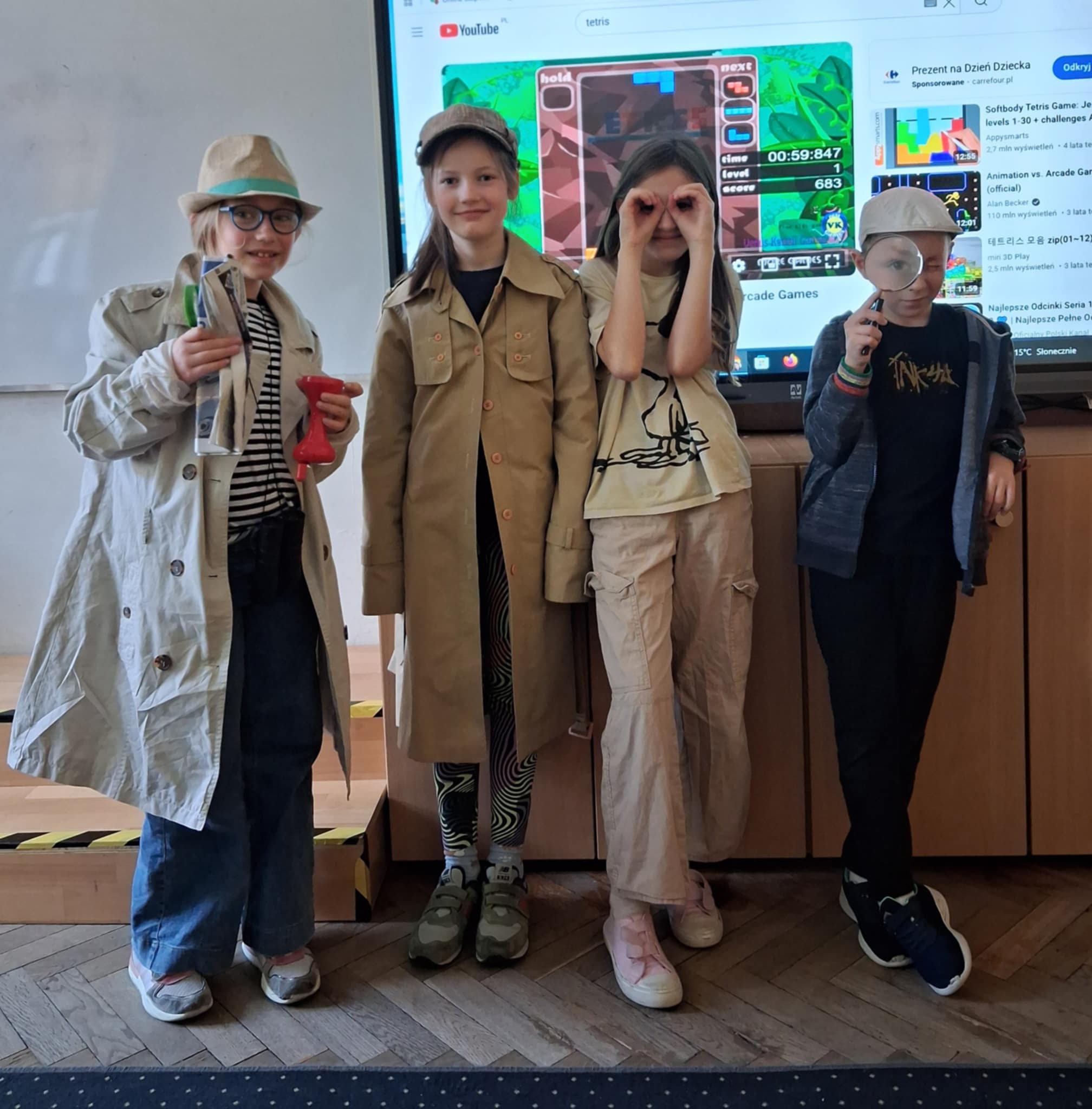 Na tle ekranu, w klasie stoi czworo dzieci. Ubrane sa w płaszcze, bluzy- przebrane są za detektywa Pozytywkę. Trzy osoby mają na głowach czapki, a w rękach trzymaja przedmioty, które kojarąa się z pracą detektywa.