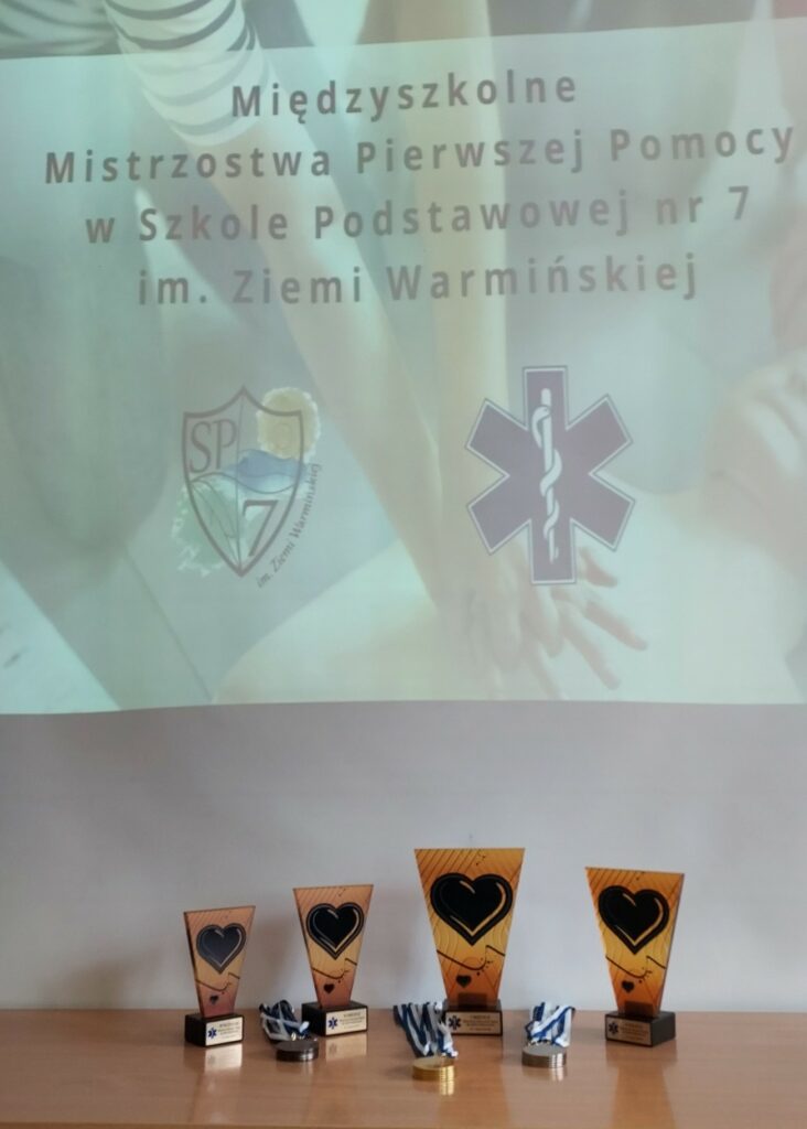 Zdjęcie przedstawia cztery statuetki ustawione obok siebie kropka Obok leżą medale brązowy złoty srebrny statuetki stoją na tle plakatu z napisem międzyszkolnym mistrzostwa pierwszej pomocy w Szkole Podstawowej nr 7 im ziemi Warmińskiej. Pod napisem w nie logo szkoły SP 7 oraz logo pierwszej pomocy medycznej.