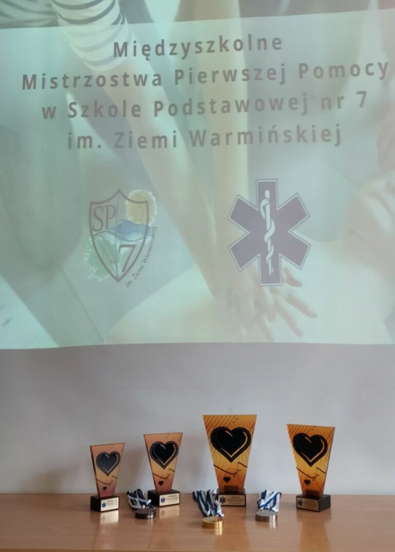 Zdjęcie przedstawia cztery statuetki ustawione obok siebie kropka Obok leżą medale brązowy złoty srebrny statuetki stoją na tle plakatu z napisem międzyszkolnym mistrzostwa pierwszej pomocy w Szkole Podstawowej nr 7 im ziemi Warmińskiej. Pod napisem w nie logo szkoły SP 7 oraz logo pierwszej pomocy medycznej.