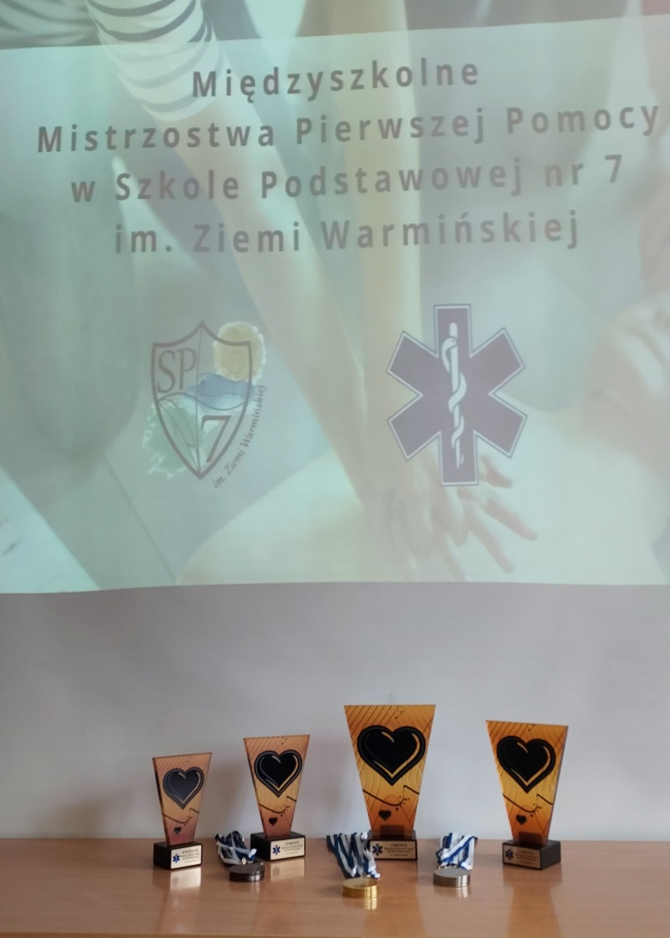 Zdjęcie przedstawia cztery statuetki ustawione obok siebie kropka Obok leżą medale brązowy złoty srebrny statuetki stoją na tle plakatu z napisem międzyszkolnym mistrzostwa pierwszej pomocy w Szkole Podstawowej nr 7 im ziemi Warmińskiej. Pod napisem w nie logo szkoły SP 7 oraz logo pierwszej pomocy medycznej.