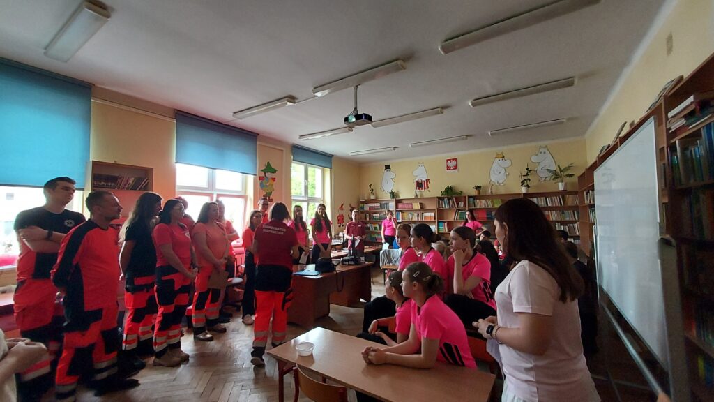 Zdjęcie przedstawia salę w której znajdują się młodzież Chłopcy i dziewczęta oraz ratownicy medyczni Mężczyźni i kobiety. Młodzież siedzi przy stolikach kropka ratownicy stoją naprzeciwko młodzieży kropka pośrodku między dwiema grupami stoi kobieta ratownik tyłem do fotografa. W tle widać regały z książkami grafiki Muminków godło rzutnik.