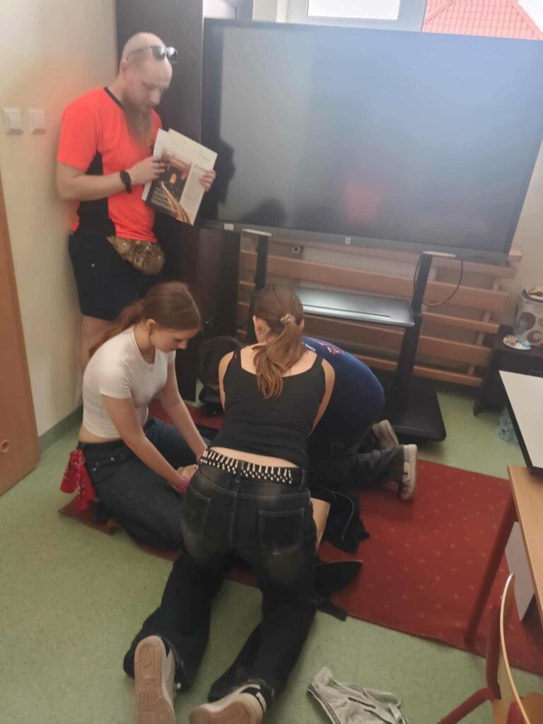 Zdjęcie przedstawia trzy osoby klęczące na kolanach pomiędzy nimi widać fantom na którym grupa wykonuje zadanie. Nad nimi stoi Medyk. Ma na głowie okulary w rękach trzeba podkładkę. Obok nich znajduje się duży telewizor stojący na statywie.