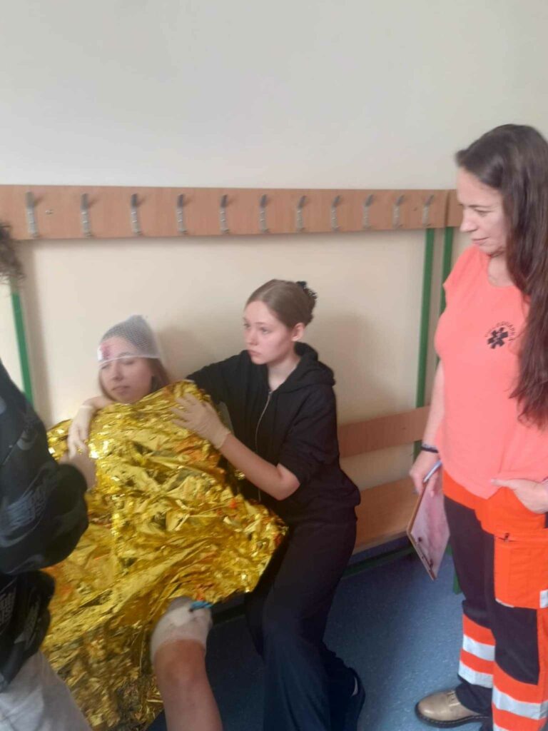 Zdjęcie przedstawia dziewczynę pół siedzącą na ławce opatuloną kocem medycznym na głowie ma założoną siatkę pod siatką widać opatrunek oraz zakrwawione czoło. Jest podtrzymywana przez dziewczynę. Obok dziewcząt stoi ratownik medyczny kobieta.