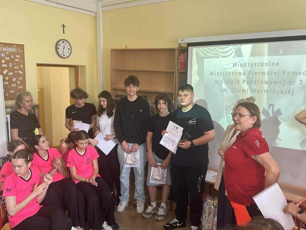 Zdjęcie przedstawia grupę chłopców i dziewcząt stojących na tle plakatu mówiącego o mistrzostwach w pierwszej pomocy medycznej. Grupa młodzieży trzyma w rękach dyplomy oraz z reklamówki z upominkami. Obok nich widać kobietę w czerwonej koszulce. Przed nimi siedzą dziewczęta ubrane w różowe koszulki czarne spodnie kropka Z drugiej strony grupy również stoi kobieta oparta o ścianę. Grupa stoi przy drzwiach które są otwarte. Nad drzwiami widać zegar