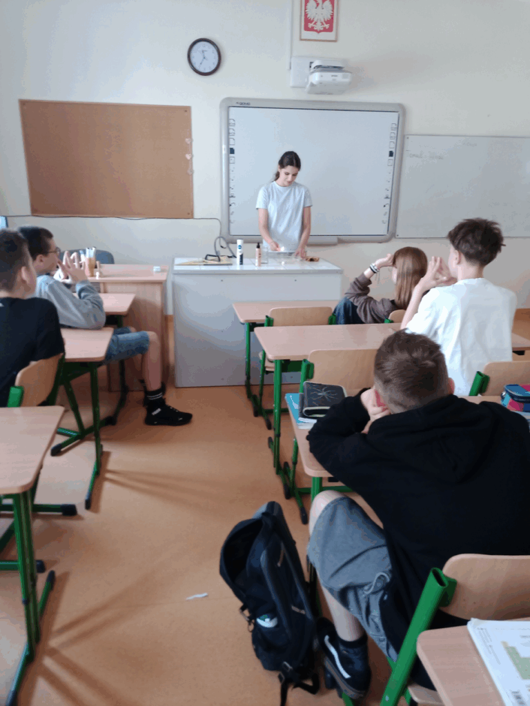 klasa lekcyjna 5 uczniów siedzi w ławkach i obserwuje koleżankę która za stołem laboratoryjnym wykonuje doświadczenie przy użyciu dezodorantu , wody i świeczki.