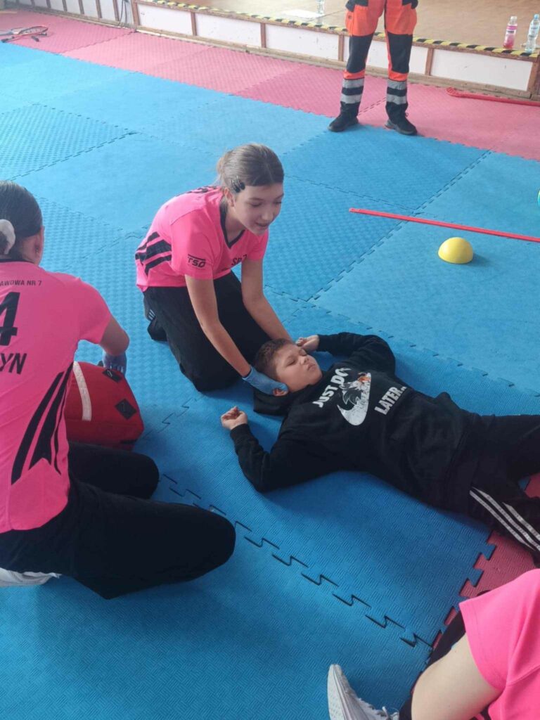 Zdjęcie przedstawia chłopca leżącego na niebieskiej macie z rozłożonymi rękoma do góry. Obok Siedzą dwie dziewczyny z czego jedno widać tylko fragment ramienia druga z odwrócona tyłem do fotografa. Trzecia dziewczyna podtrzymuje głowę leżącego chłopca kropka Z tyłu widać nogi ubrane w pomarańczowe spodnie.