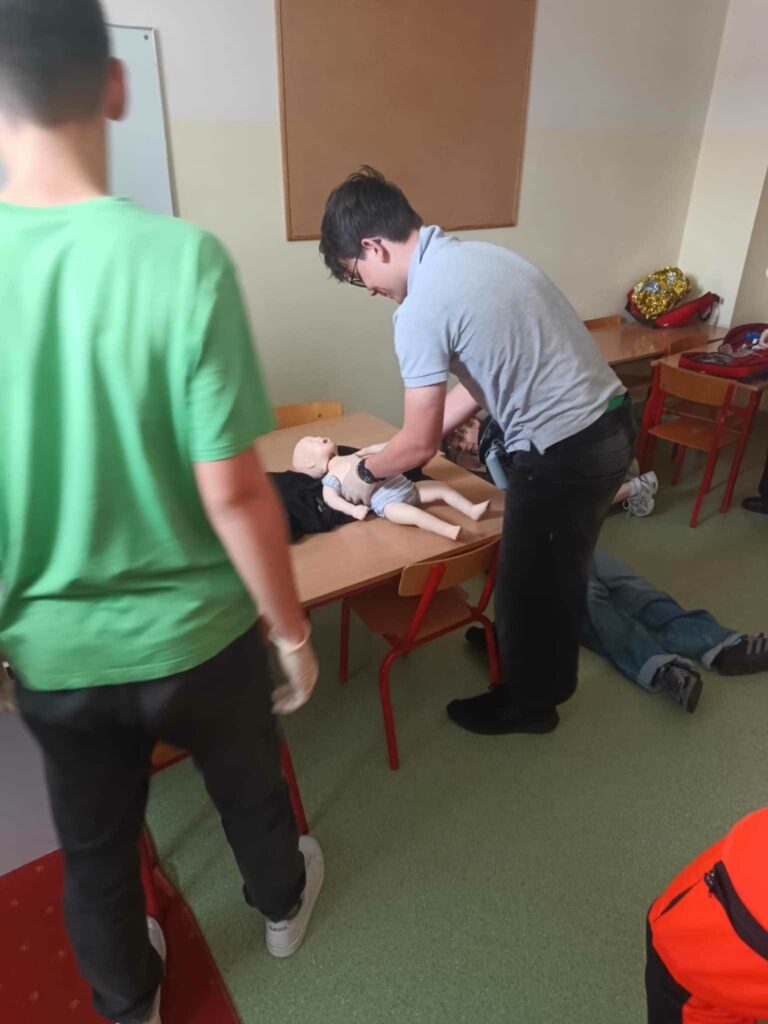 Zdjęcie przedstawia chłopca stojącego przy ławkach na którym leży Phantom małego dziecka Chłopiec wykonuje resuscytację. Tyłem do fotografa stoi inny chłopiec w zielonej koszulce.