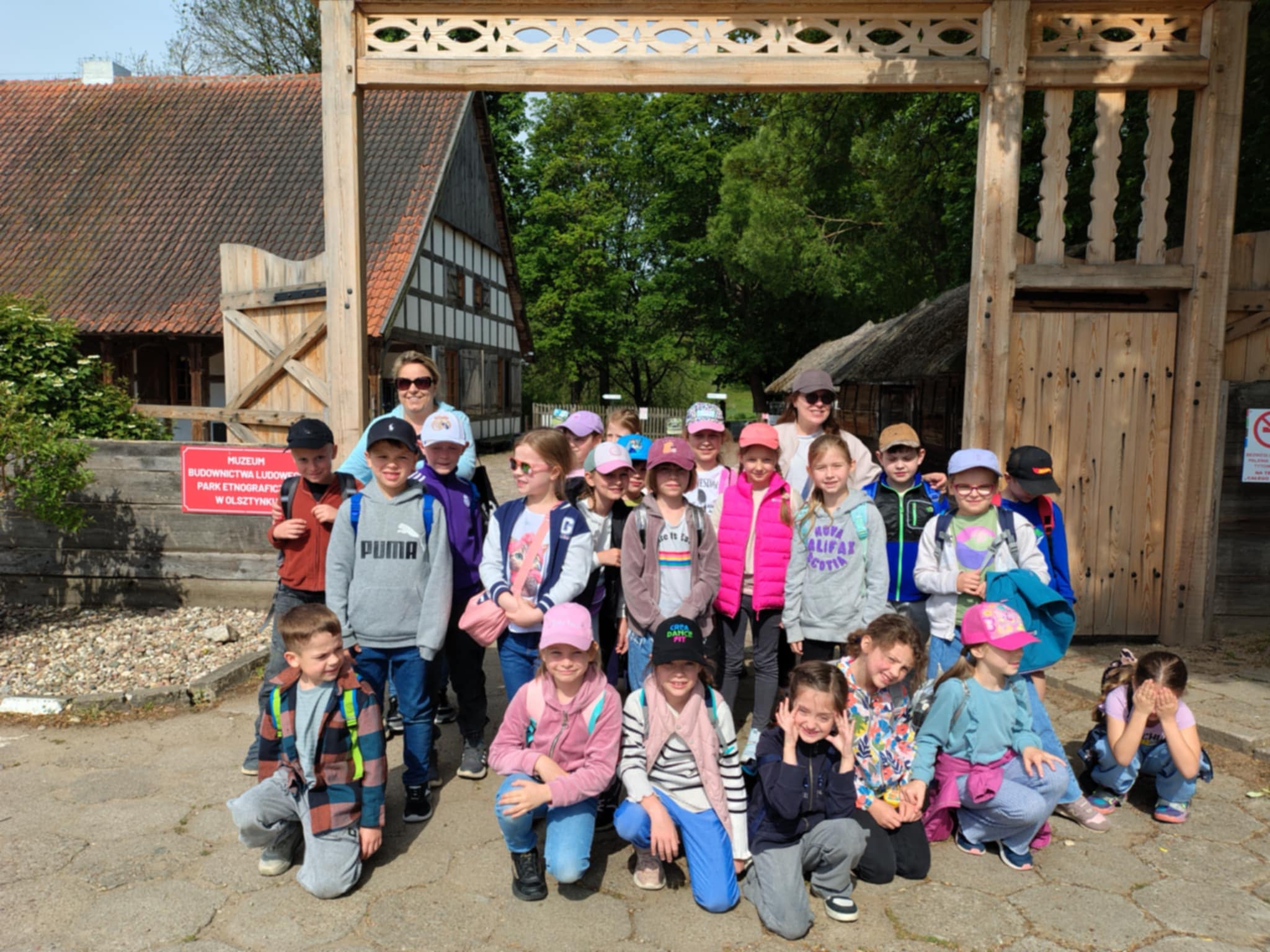 Grupa dzieci na wycieczce szkolnej, pozuje do zdjęcia przed dużą, drewnianą bramą. Za nimi widać tradycyjne, drewniane zabudowania. Dzieci zwiedzają skansen. Część dzieci stoi, pozostałe kucają przed nimi.
