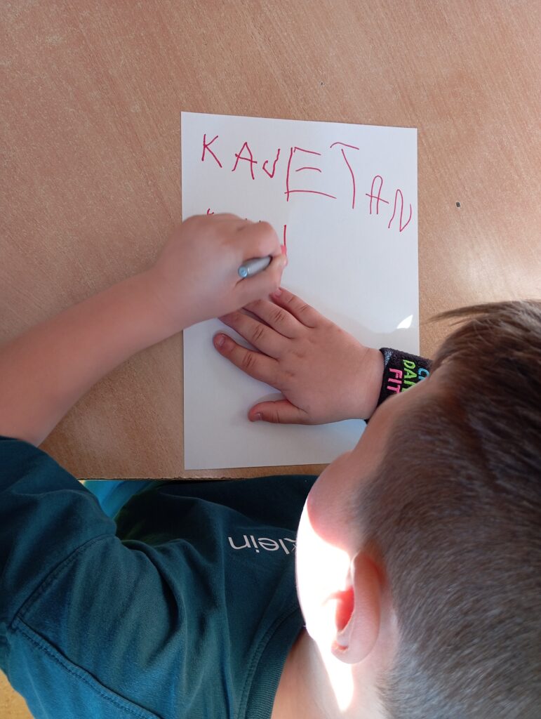 Na zdjęciu widać chłopca siedzącego przy stoliku. Chłopiec trzyma w lewej ręku marker czerwony i pisze swoje imię Kajetan na białej kartce. Pismo jest niekształtne, ponieważ uczeń jest praworęczny.