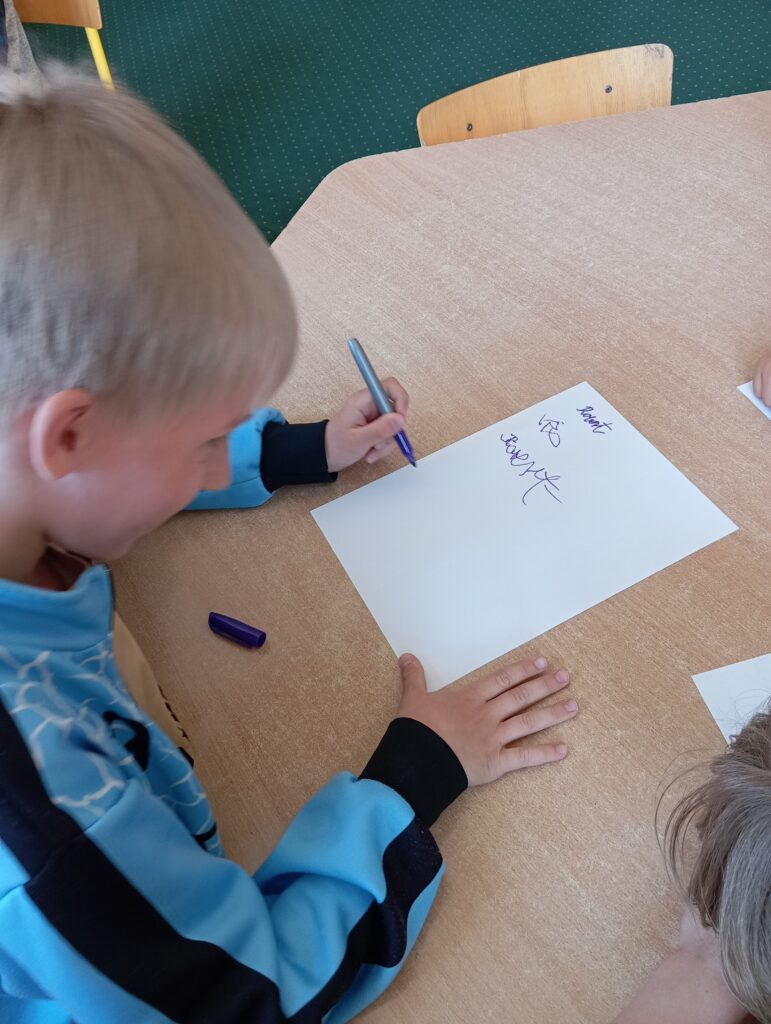 Na zdjęciu widać chłopca siedzącego przy stoliku. Uczeń jest w niebieskiej bluzie. W lewej ręku trzyma niebieski markier i pisze swoje imię Robert na białej kartce. Imię jest niekształtne ponieważ uczeń jest praworęczny.