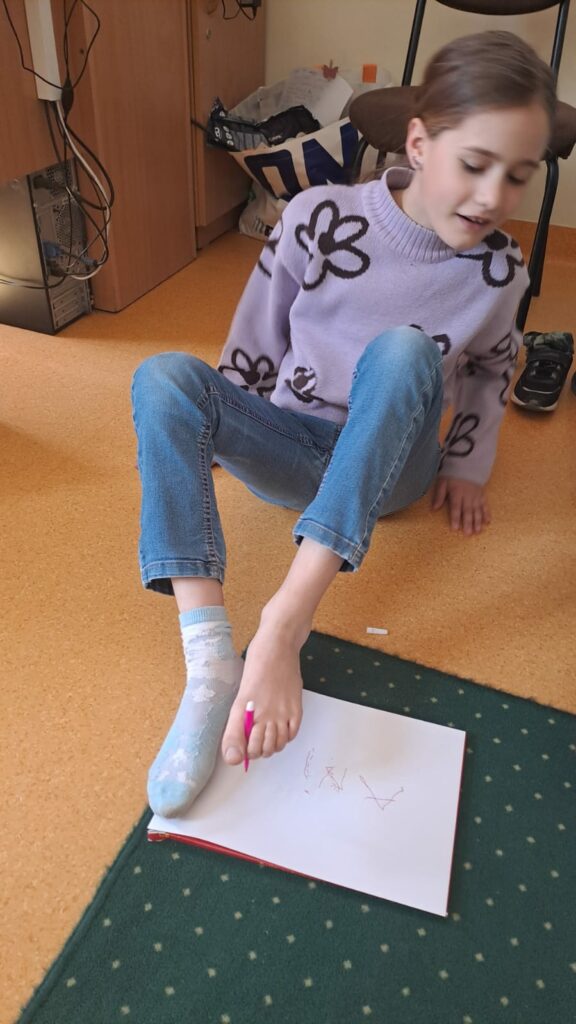 Na zdjęciu widać dziewczynkę, która siedzi na podłodze. W lewej stopie- palcach trzyma różowy mazak i pisze swoje imię Aniela na białej kartce.