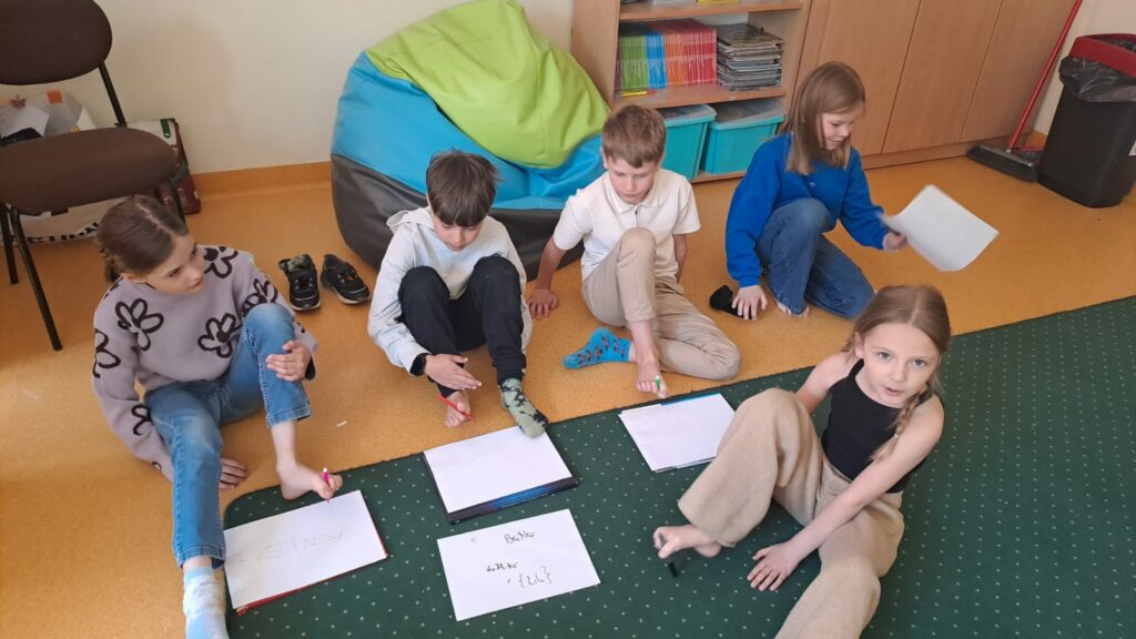 Na zdjęciu widać 3 dziewczynki, 2 chłopców, którzy siedzą na podłodze. W palcach u stóp trzymają mazaki i piszą na białych kartkach.