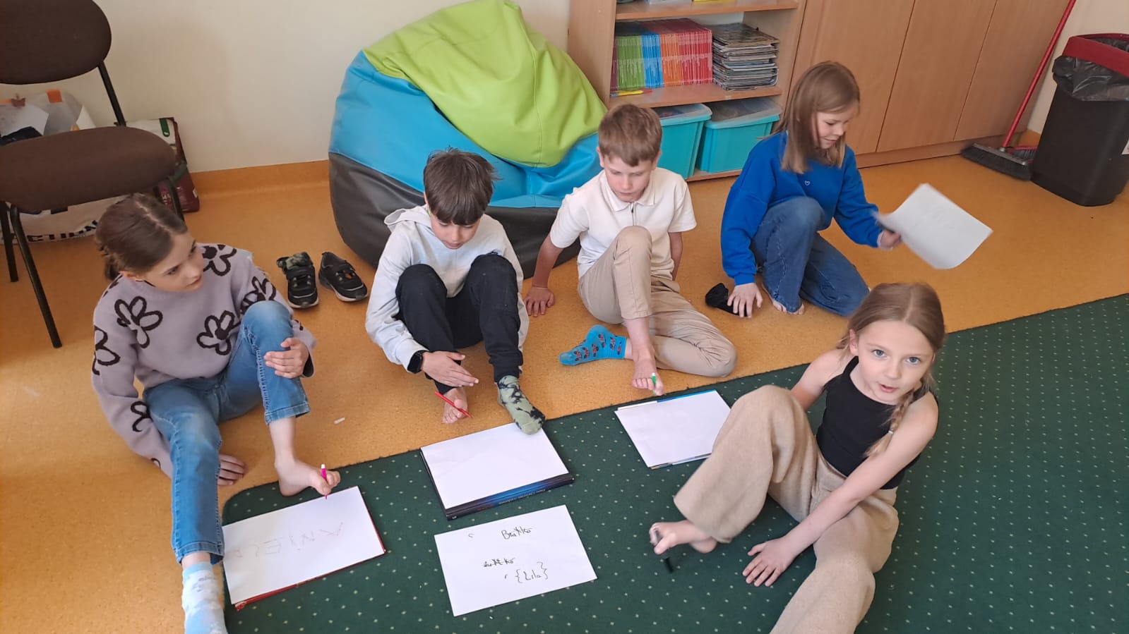 Na zdjęciu widać 3 dziewczynki, 2 chłopców, którzy siedzą na podłodze. W palcach u stóp trzymają mazaki i piszą na białych kartkach.