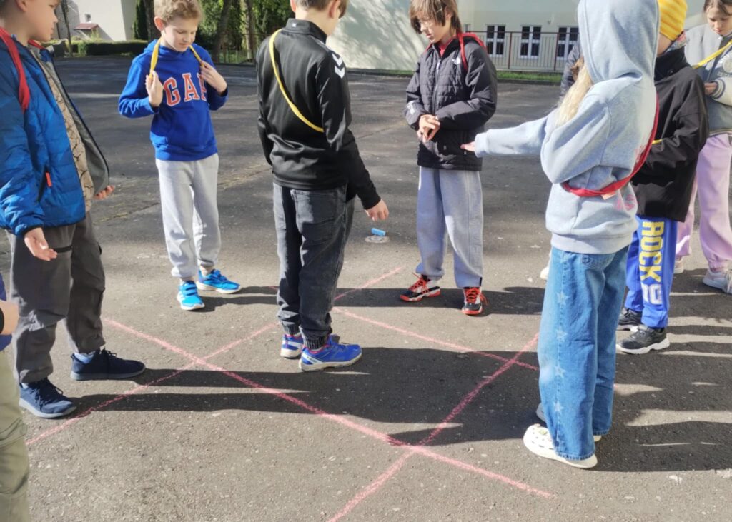 Na zdjęciu widzimy grupę dzieci biorących udział grze na świeżym powietrzu, na boisku szkolnym. Dzieci stoją w kręgu wokół narysowanej na asfalcie figury z różowej kredy – jest to duży kwadrat podzielony na mniejsze pola. Kredowy kwadrat może służyć jako plansza do gry terenowej, np. „kółko i krzyżyk” Każde dziecko ma zawieszony na szyi kolorowy sznurek lub wstążkę (czerwony lub żółty), co może sugerować podział na zespoły. Dzieci mają luźne, sportowe stroje, odpowiednie do aktywności na dworze. W tle widać budynek szkolny, co potwierdza, że scena ma miejsce na terenie szkoły. Panuje słoneczna pogoda – dzieci rzucają wyraźne cienie na asfalt. Atmosfera wydaje się być pełna zaangażowania i współpracy.