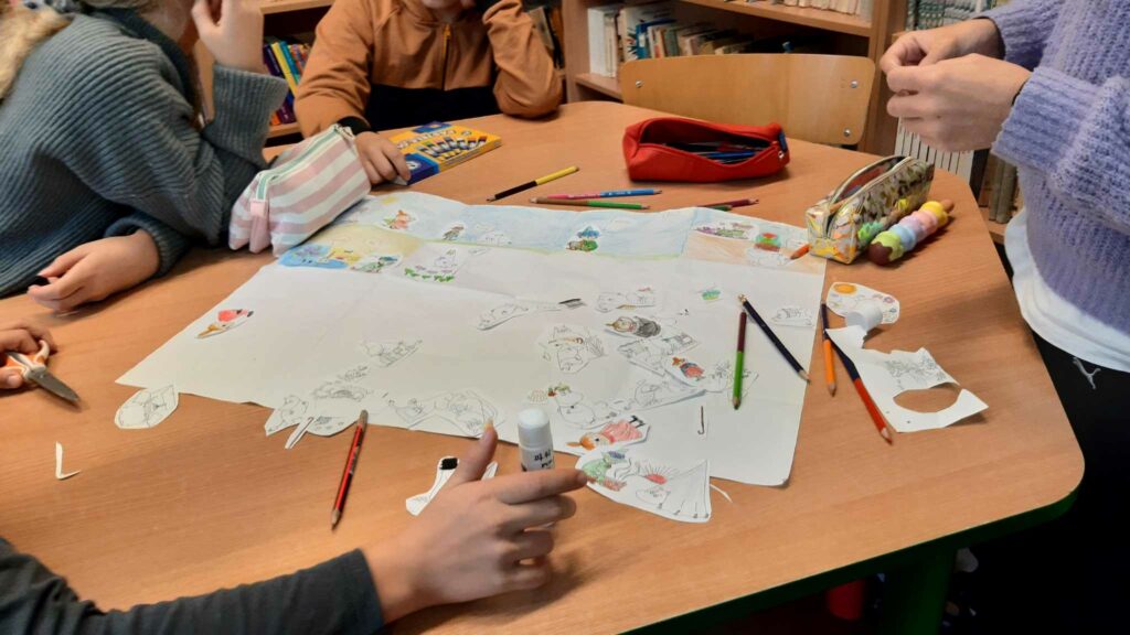 Zdjęcie przedstawia stół na którym znajduje się Biała kartka papieru na niej rysunki okienka komiksu. Wokół kartki i na kartce znajdują się przybory papiernicze kredki ołówki kleje nożyczki powycinane fragmenty postaci przedstawiające Muminki i innych bohaterów z tej bajki. Na stole widać również piórniki oraz dłonie dzieci które wykonują pracę plastyczną w tle widać książki na półkach.