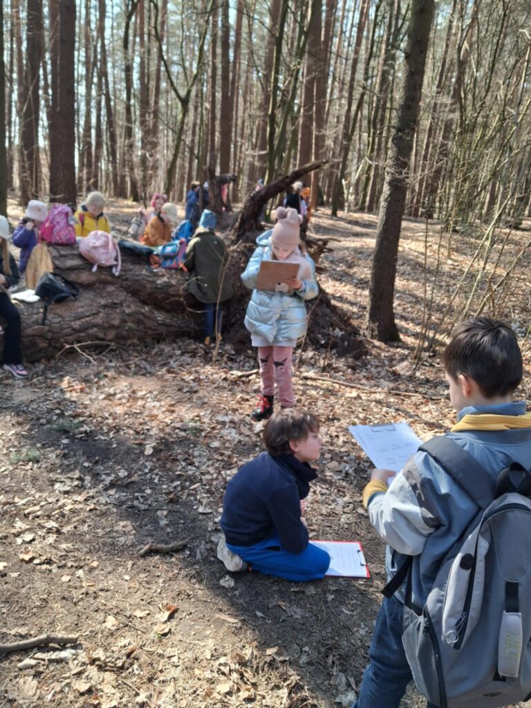 Dzieci uczestniczą w lekcji przyrody na świeżym powietrzu, w lesie. Część dzieci stoi na ścieżce leśnej, niektóre stoją przy pniu drzewa i notują obserwacje w zeszytach. Uczniowie są skupieni i zaangażowani w pracę. W tle widać drzewa i naturalne otoczenie, które sprzyja nauce poprzez bezpośredni kontakt z przyrodą.