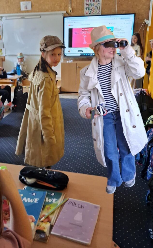 W sali klasowej, na pierwszym planie widać dwie dziewczynki. Obie ubrane sa w płaszcze, a na głowach maja kapelusze. Jedna patrzy przez lornetkę, a w reku trzyma zwinięta gazetę. Druga rozgląda się dookoła. Dziewczynki naśladują zachowanie detektywa.