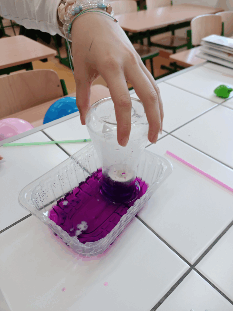 Na stole laboratoryjnym widać kolbę zanurzoną w wodzie o kolorze fioletowym. W kolbie umieszczona jest świeczka która z braku powierza gaśnie a poziom wody w kolbie się podnosi.