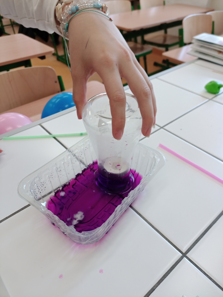 Na stole laboratoryjnym widać kolbę zanurzoną w wodzie o kolorze fioletowym. W kolbie umieszczona jest świeczka która z braku powierza gaśnie a poziom wody w kolbie się podnosi.