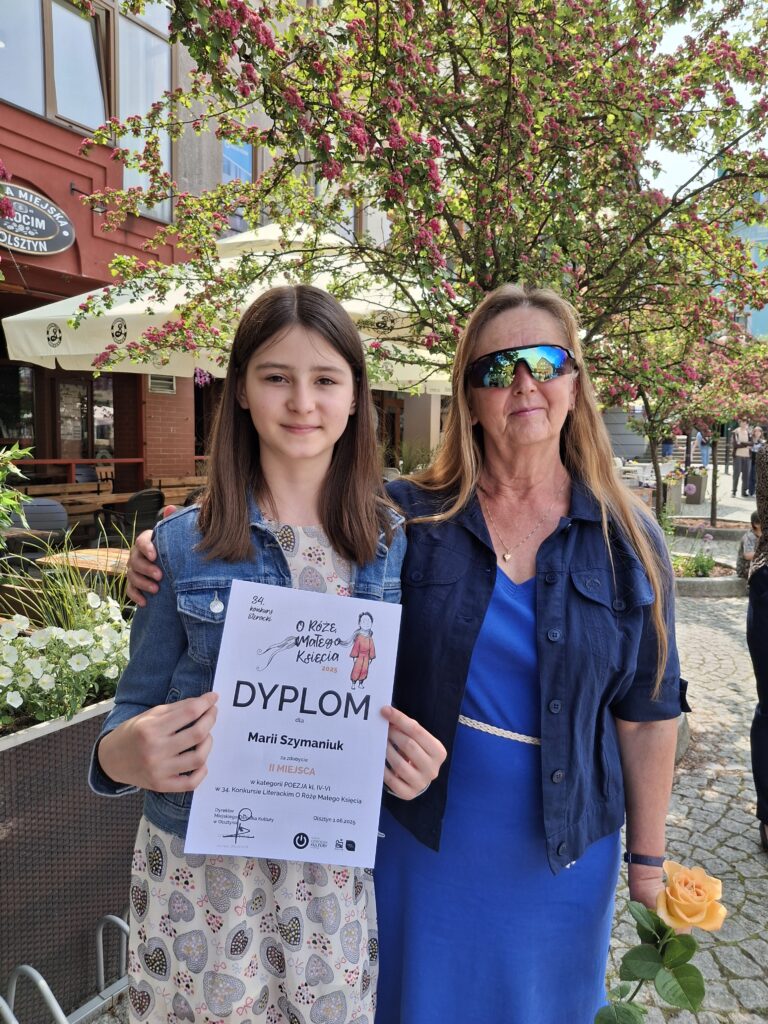 zdjęcie :po lewej uczennica w niebieskiej bluzie, po prawej nauczycielka w granatowej bluzie i niebieskiej sukience.Dziewczynka trzyma w dłoniach dyplom kobieta żółtą różę