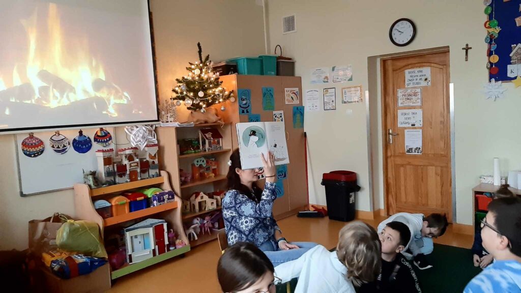 Zdjęcie przedstawia salę w której znajdują się dzieci siedzące na podłodze przed nauczycielem kobietą która trzyma w dłoni podniesioną nad głowę książkę otwartą w której są postacie z Muminków. W tle widać fragment ekranu na którym Płonie ognisko. Obok ekranu znajdują znajduje się półka na której stoi choinka. A wokół dzieci znajdują się półki z zabawkami widać drzwi oklejone kolorowymi wydrukami nad drzwiami wisi zegar obok wisi krzyż.