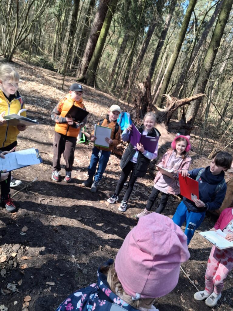 Dzieci stoja w kole. Kazdy ma kartkę z notatkami. Dziela sie efektami swojej pracy. Zdjecie zrobione jest w lesie.