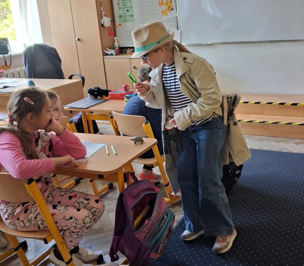 W sali szkolnej, na pierwszym planie stoi bokiem dziewczynka ubrana w płaszcz i kapelusz. Patrzy przez lupę- naśladuje zachowanie detektywa. Stoi przy ławce szkolnej, przy której siedzą dziewczynki.