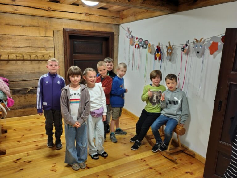 Zdjęcie przedstawia grupę dzieci uczestniczących w warsztatach w skansenie w Olsztynku, gdzie poznają proces powstawania swetrów. Dzieci stoją i siedzą w pomieszczeniu o drewnianych ścianach i podłodze, a za nimi widoczna jest dekoracja z kolorowych masek i wstążek. Uczestnicy wyglądają na zadowolonych i zaangażowanych, co świadczy o przyjaznej atmosferze zajęć edukacyjnych.