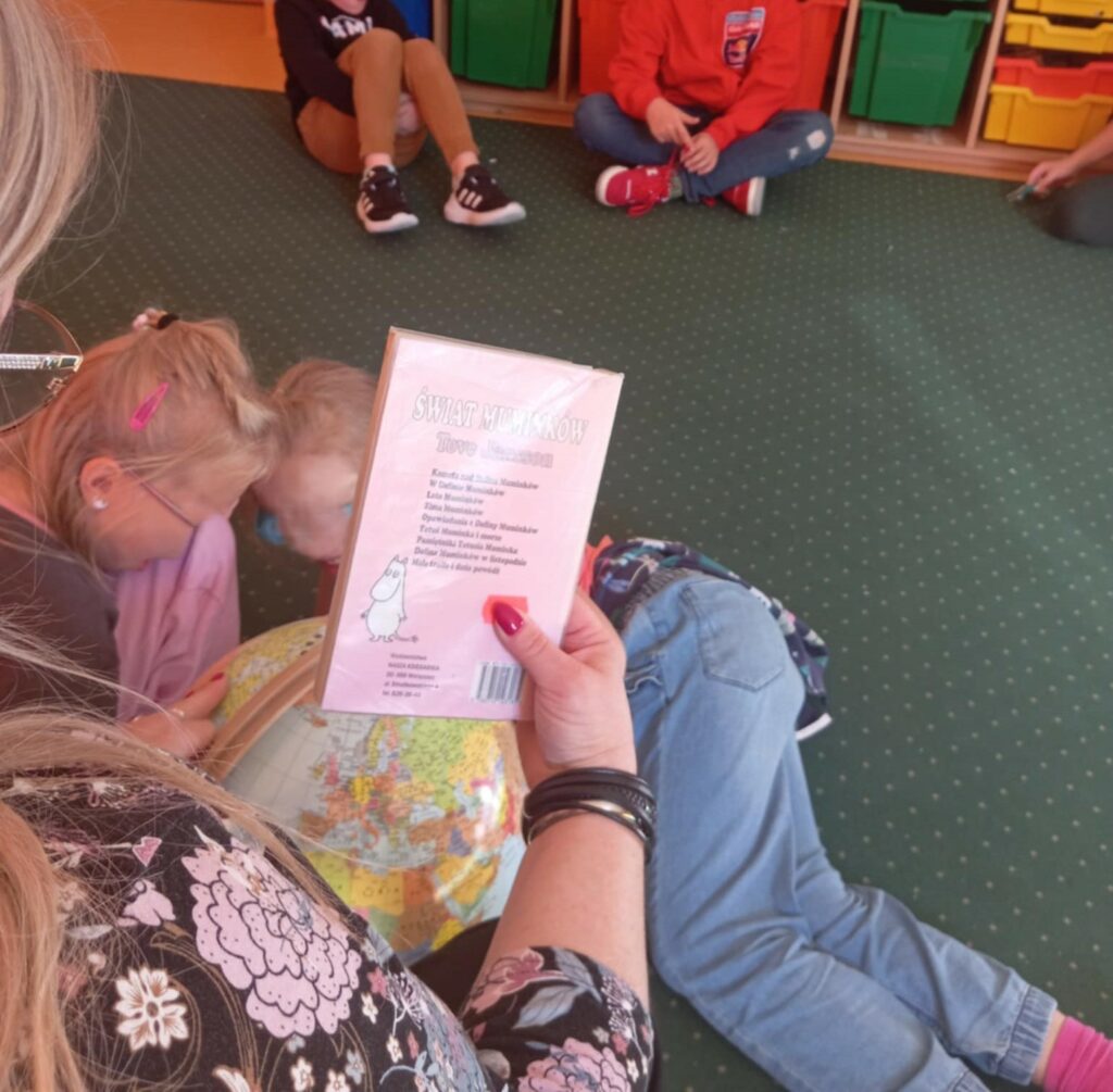 Zdjęcie przedstawia fragment kobiety stojącej tyłem do fotografa trzymającej w dłoni książkę świat Muminków a drugą dłoń opiera na globusie wskazując na nim coś. Wokół globusu obok kobiety znajdują się dzieci przyglądające się globusowi. W tle widać dzieci siedzące na dywanie po turecku.