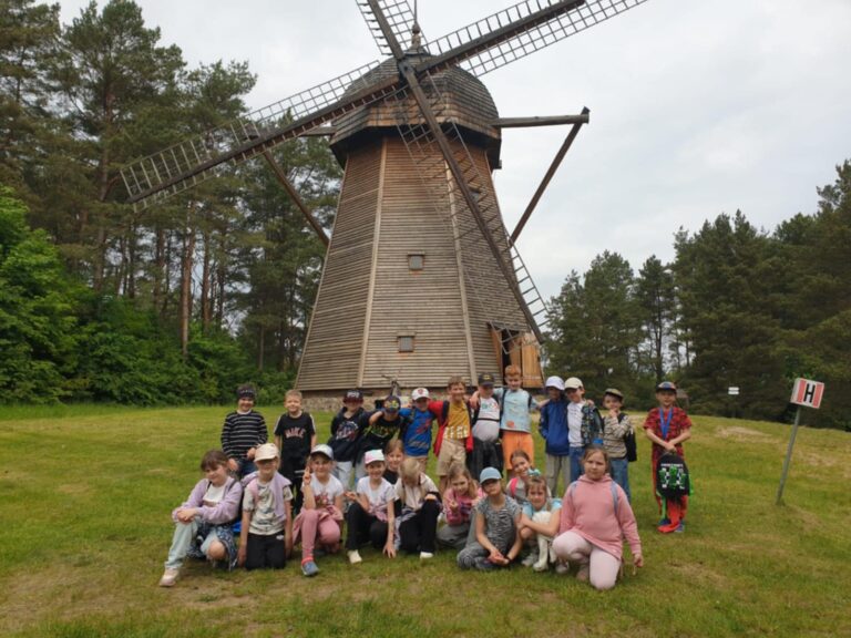 Dzieci z klasy 1A odwiedzily skansen w Olsztynku i zrobiła sobie pamiątkowe zdjęcie przed dużym drewnianym wiatrakiem. Dzieci ubrane są kolorowo i uśmiechnięte, stoją i siedzą na zielonej trawie. W tle widać las oraz fragment tablicy z literą „H”. Wycieczka odbyła się w pogodny dzień i z pewnością była ciekawą przygodą edukacyjną.