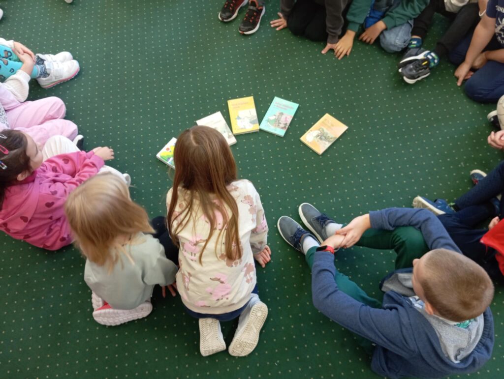 Zdjęcie przedstawia dzieci siedzące w kręgu większość jest zwrócona tyłem do fotografa kropka W kręgu leżą rozłożone książki w liczbie 5 przedstawiające Muminki.