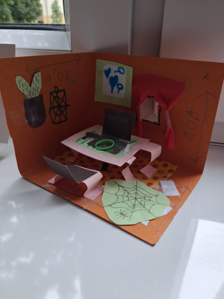 Fotografia przedstawia makietę biura detektywistycznego, wykonaną z papieru. Jest stolik i krzesełka. Wykonane sa okna z firankami.