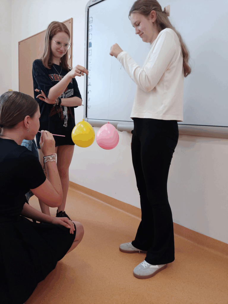 Na zdjęci stoją dwie dziewczynki które trzymają balon zawieszone na nitkach. Przed nimi kuca koleżanka która za pomocą słomki przez którą dmucha oddziela balony od siebie