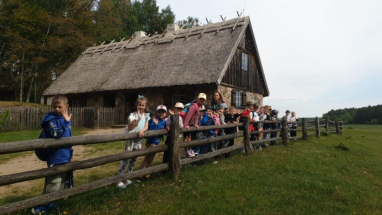 Na zdjęciu widzimy grupę dzieci stojącą przy drewnianym płocie, przed zabytkową chatą krytą strzechą w skansenie w Olsztynku. Dzieci ubrane są na sportowo, wiele z nich ma plecaki i czapki, co sugeruje, że są na wycieczce szkolnej. W tle widać drewniany dom oraz otoczenie pełne zieleni i drzew, co oddaje klimat wiejskiego muzeum na świeżym powietrzu. Dzieci uśmiechają się i machają do aparatu, tworząc radosną atmosferę.