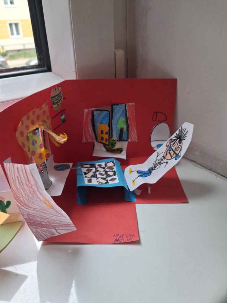 Fotografia przedstawia makietę biura detektywistycznego, wykonaną z papieru. Jest stolik i krzesełka. Wykonane sa okna z firankami.