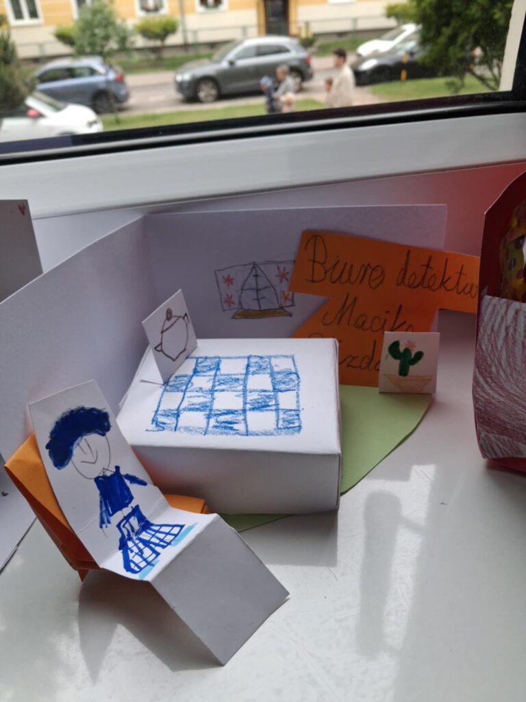 Fotografia przedstawia makietę biura detektywistycznego, wykonaną z papieru. Jest stolik i krzesełka. Wykonane sa okna z firankami.